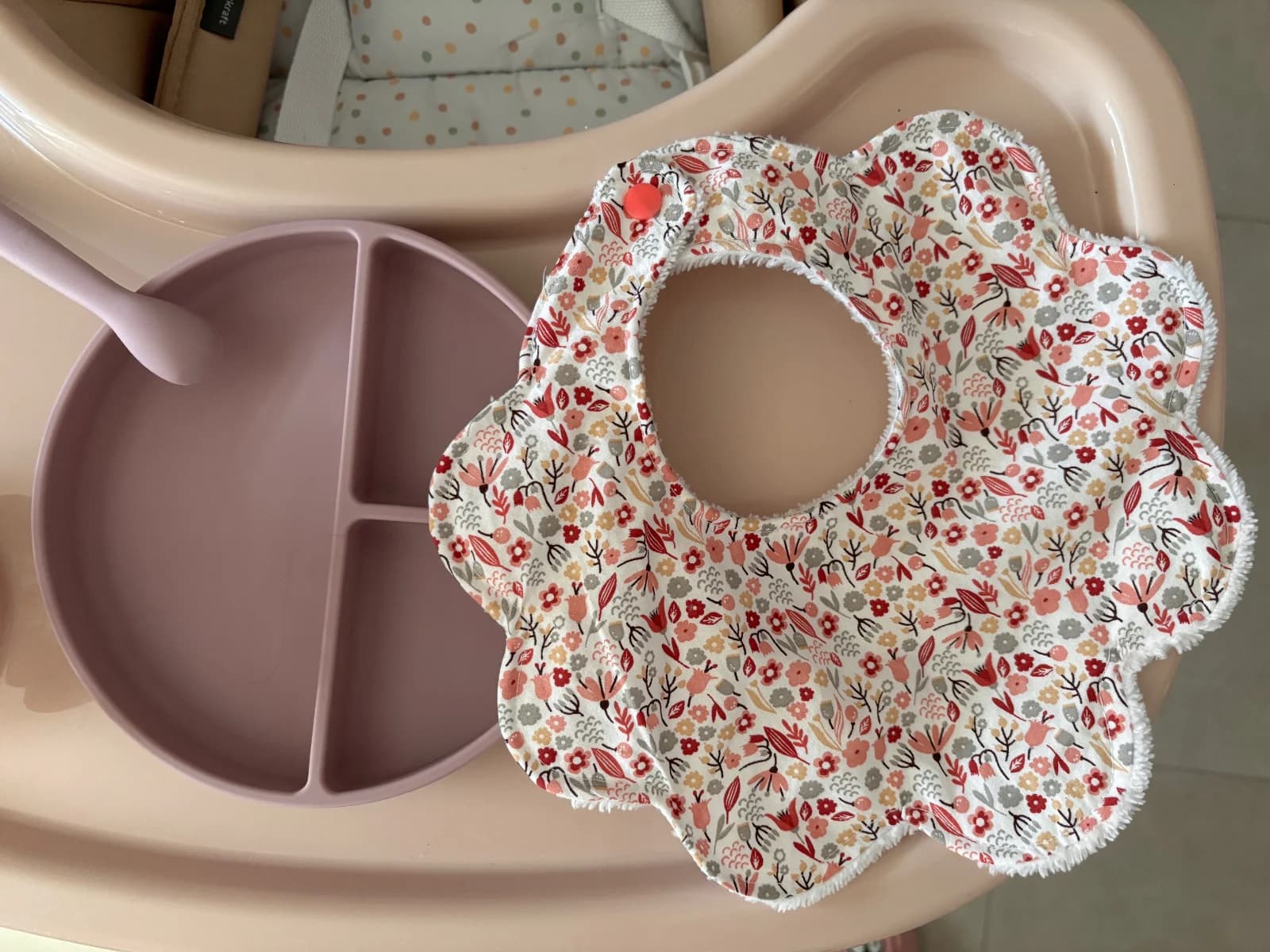 Bavoir en forme de fleur à motif floral rose et terracotta posé près d'une assiette pour bébé.