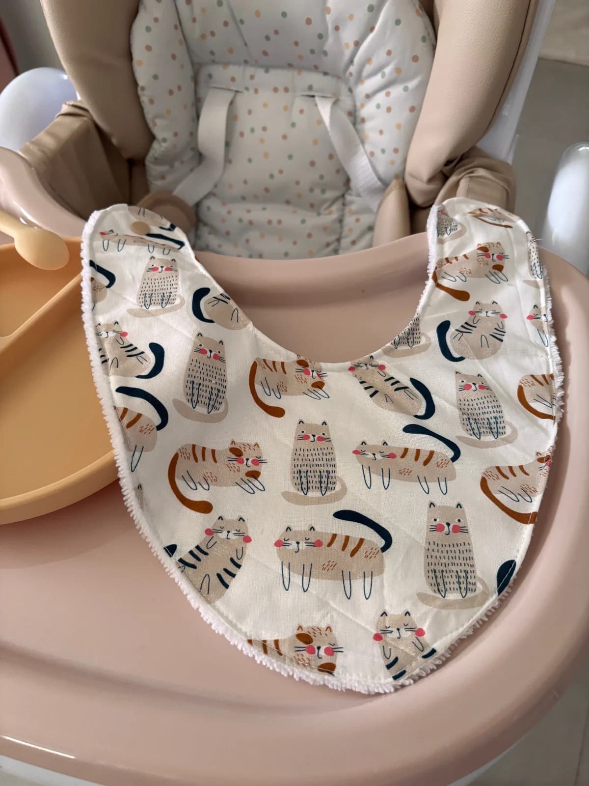 Bavoir bandana blanc à motifs de chats beiges et rayés posé à plat dans une chaise haute.