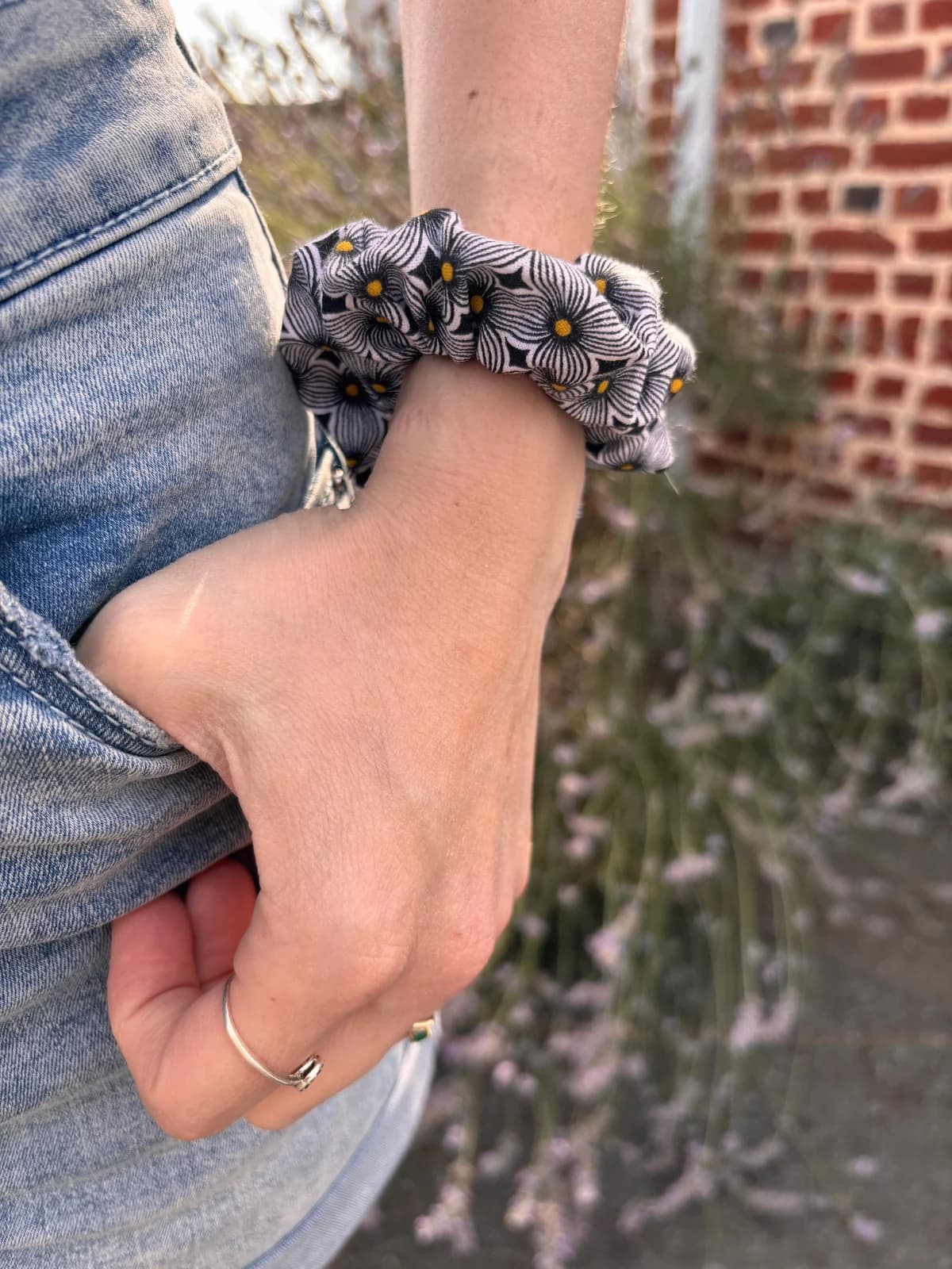 Chouchou à motif floral noir, blanc et jaune porté au poignet, main dans la poche d'un jean.